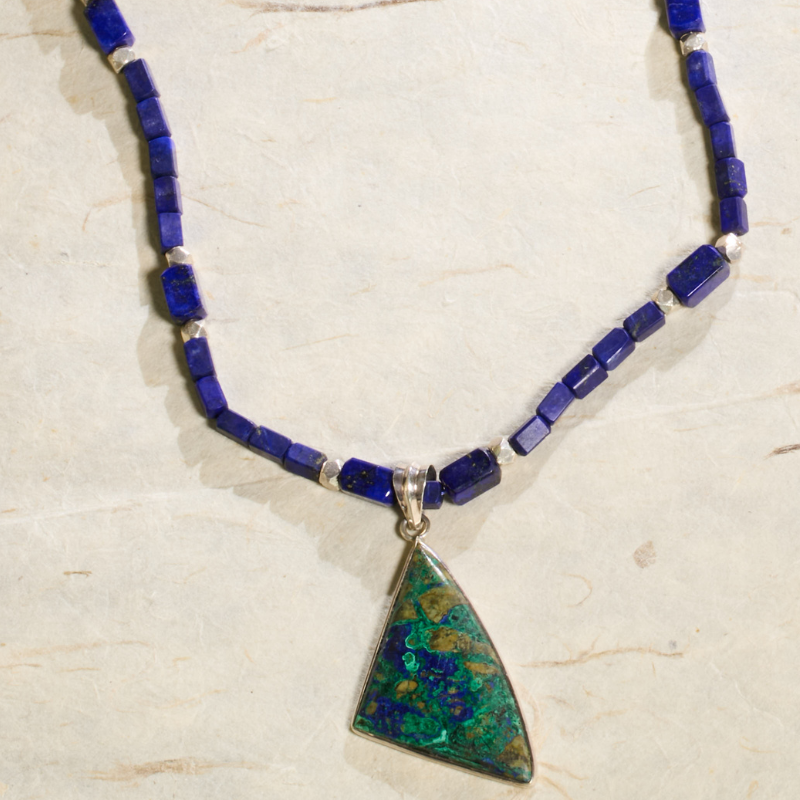 Top-down studio shot of Lapis Lazuli beaded necklace with a triangular green Malachite pendant on a textured cream background with natural elements.